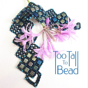 以下が含まれることがあります： 青、緑、金色の色合いの幾何学模様のビーズブレスレット。ブレスレットは小さな正方形のビーズで作られており、ジグザグ模様に配置されています。ブレスレットは、紫色の花が咲いた白い背景に飾られています。