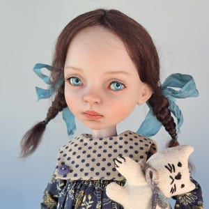 Pu&ograve; includere: Una bambola fatta a mano con trecce marroni adornate da nastri blu. La bambola ha occhi blu, guance rosee e un vestito a fantasia floreale. Tiene in mano un piccolo giocattolo a forma di gatto di peluche. L'abito della bambola ha un colletto a pois.