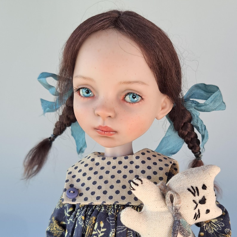 Pu&ograve; includere: Una bambola fatta a mano con trecce marroni adornate da nastri blu. La bambola ha occhi blu, guance rosee e un vestito a fantasia floreale. Tiene in mano un piccolo giocattolo a forma di gatto di peluche. L'abito della bambola ha un colletto a pois.