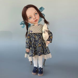 Pu&ograve; includere: Bambola fatta a mano con occhi blu, capelli castani a trecce e un vestito a fantasia floreale. La bambola tiene in mano un peluche e indossa calzamaglia bianca e scarpe blu. L'abito ha un colletto a pois e un bordo in pizzo.