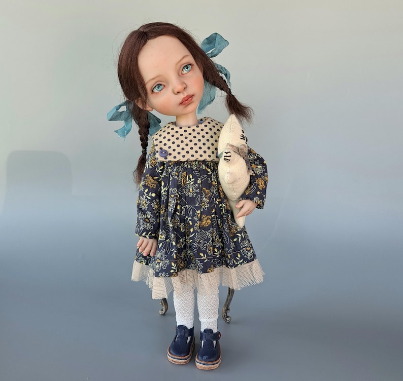 Pu&ograve; includere: Bambola fatta a mano con occhi blu, capelli castani a trecce e un vestito a fantasia floreale. La bambola tiene in mano un peluche e indossa calzamaglia bianca e scarpe blu. L'abito ha un colletto a pois e un bordo in pizzo.