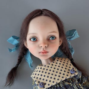 Pu&ograve; includere: Una bambola artigianale con un'espressione serena, occhi blu e capelli castani intrecciati adornati da nastri blu. La bambola indossa un vestito con colletto a pois e due bottoni blu. Il viso della bambola ha un aspetto delicato.