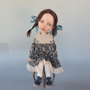 Pu&ograve; includere: Bambola artigianale con trecce castane ornate da nastri blu. La bambola indossa un vestito floreale blu navy con colletto a pois, finiture in pizzo bianco e scarpe blu navy. La bambola &egrave; seduta su una sedia decorativa.
