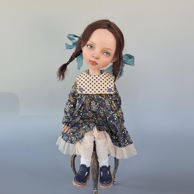 Pu&ograve; includere: Bambola artigianale con trecce castane ornate da nastri blu. La bambola indossa un vestito floreale blu navy con colletto a pois, finiture in pizzo bianco e scarpe blu navy. La bambola &egrave; seduta su una sedia decorativa.