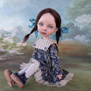 Pu&ograve; includere: Una bambola artigianale con trecce marroni adornate da nastri blu. La bambola indossa un vestito floreale blu navy con colletto a pois bianchi e bordatura in pizzo bianco. La bambola ha gli occhi blu e indossa scarpe blu navy.