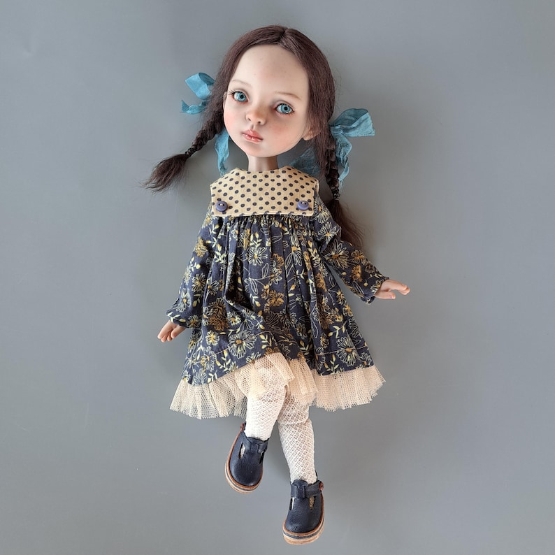 Pu&ograve; includere: Una bambola fatta a mano con occhi blu, trecce castane e un vestito con stampa floreale. L'abito ha un bordo in pizzo beige e un colletto a pois. La bambola indossa calze bianche e scarpe blu navy.