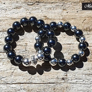 May include: Two bracelets, one black and one silver, with round beads. The bracelets are linked together by a silver charm.