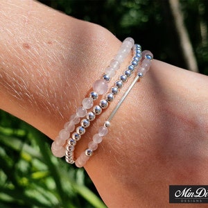 May include: A set of three bracelets featuring pink and silver beads. The bracelets are stacked on a person's wrist. The bracelets are made with small, round beads and a silver bar.