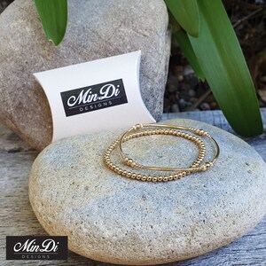 May include: Two gold beaded bracelets, one with larger beads and one with smaller beads, are stacked on top of each other. The bracelets are on a light brown surface. The bracelets are packaged in a white pillow box with the text "Min Di Designs" printed on the front.