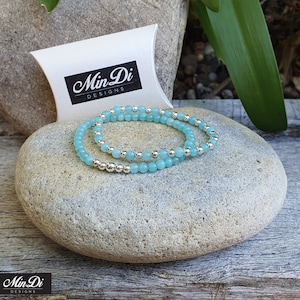 May include: Two turquoise blue beaded bracelets with silver beads, on a grey stone, with a white box behind the bracelets. The box has the text "Min Di Designs" on it.