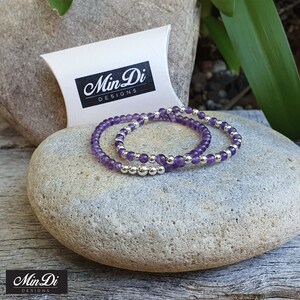 May include: Two amethyst gemstone bracelets with silver beads. The bracelets are stacked on top of each other and are on a grey stone. The bracelets are packaged in a white pillow box with the text "Min Di Designs" printed on the box.