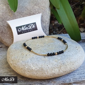 May include: A gold bracelet with black beads and gold accents. The bracelet is on a gray rock.