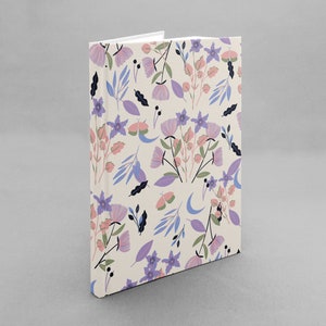 Op de afbeelding: Een crèmekleurig notitieboek met een bloemenpatroon met roze, paarse en blauwe bloemen, bladeren en halve manen.