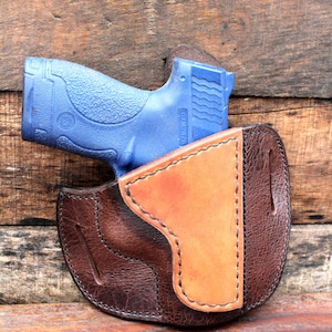 May include: A brown leather holster with a tan leather lining. A blue handgun is partially inserted into the holster.