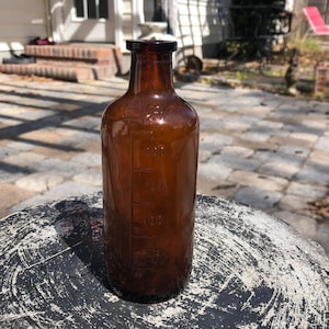 Könnte beinhalten: Eine braune Glasflasche mit einer graduierten Skala, die in Millilitern gekennzeichnet ist. Die Flasche steht auf einer runden, grauen Oberfläche mit weißen Markierungen.