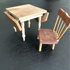 May include: A miniature wooden table with drop leaves and a matching chair. The table has light-colored legs and a natural wood finish on the tabletop. The chair has a dark brown seat and a spindle back. The table is approximately 6 inches long.