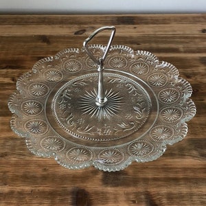 May include: A clear glass serving tray with a silver handle. The tray has a scalloped edge and a decorative pattern of circles and flowers.