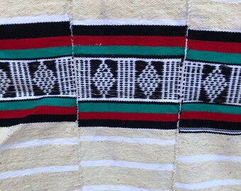 May include: A white woven fabric with horizontal stripes and a patterned border. The border features black, red, and green stripes with a geometric design.