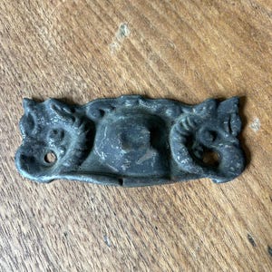 May include: A dark, antique-style metal drawer pull with ornate ram's head designs on each end. The pull has a central, slightly raised section and two screw holes. The metal has a weathered, aged appearance, suggesting it is vintage.