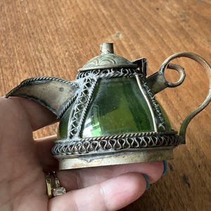 以下が含まれることがあります： 緑色のガラスの本体と複雑な金属細工のディテールを持つ、小さな銀のティーポット。ティーポットは湾曲した注ぎ口とハンドルを持っています。
