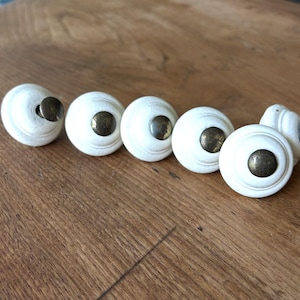 Może przedstawiać: Sześć białych, okrągłych gałek do szafek z mosiężnymi metalowymi akcentami. Gałki ułożone są w rzędzie na drewnianej powierzchni. Gałki mają lekko teksturowaną powierzchnię i klasyczny design.