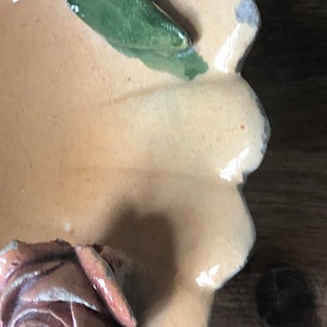 May include: A ceramic dish with a scalloped edge, decorated with a raised, light brown rose and a green leaf. The dish is glazed in a light beige color.