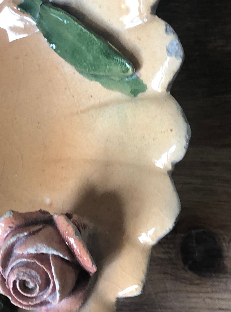 May include: A ceramic dish with a scalloped edge, decorated with a raised, light brown rose and a green leaf. The dish is glazed in a light beige color.
