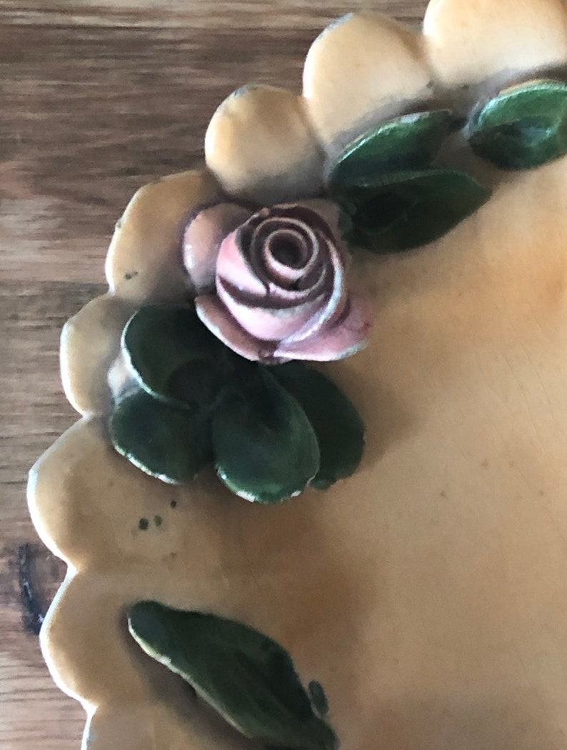May include: A cream-colored ceramic dish with a scalloped edge. The dish is decorated with a pink rose and green leaves.