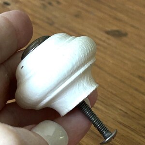 Peut inclure: Un bouton de meuble blanc et orn&eacute;, avec un accent circulaire fonc&eacute; sur le dessus. Le bouton est tenu par une main et poss&egrave;de une vis m&eacute;tallique filet&eacute;e pour la fixation. Le bouton est probablement pour un tiroir ou une armoire.