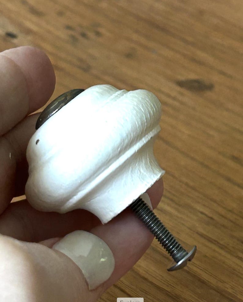 Peut inclure: Un bouton de meuble blanc et orn&eacute;, avec un accent circulaire fonc&eacute; sur le dessus. Le bouton est tenu par une main et poss&egrave;de une vis m&eacute;tallique filet&eacute;e pour la fixation. Le bouton est probablement pour un tiroir ou une armoire.