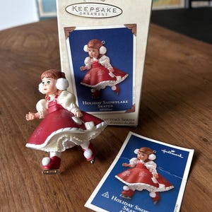 Può includere: Un ornamento Hallmark Keepsake raffigurante una pattinatrice. La statuina indossa un vestito rosso con finiture bianche e paraorecchie. La scatola e la carta mostrano anche la statuina. Il testo recita "Holiday Snowflake Skater Alexander".