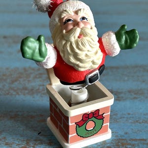 May include: A vintage Santa Claus ornament with a spring-loaded mechanism that makes him pop up from a brick chimney. The chimney is decorated with a red ribbon and a green wreath with the year 2001 printed on it.