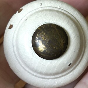 Peut inclure: Gros plan sur un bouton de meuble en bois peint en blanc avec un centre en laiton. Le bouton a un design circulaire avec une finition l&eacute;g&egrave;rement vieillie, montrant de petites zones de bois apparent. Le centre en laiton a un aspect marbr&eacute; et vieilli.