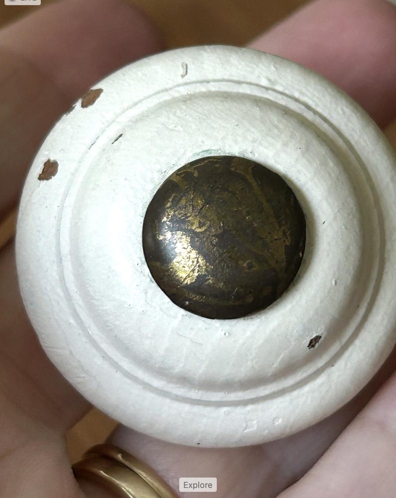 Peut inclure: Gros plan sur un bouton de meuble en bois peint en blanc avec un centre en laiton. Le bouton a un design circulaire avec une finition l&eacute;g&egrave;rement vieillie, montrant de petites zones de bois apparent. Le centre en laiton a un aspect marbr&eacute; et vieilli.