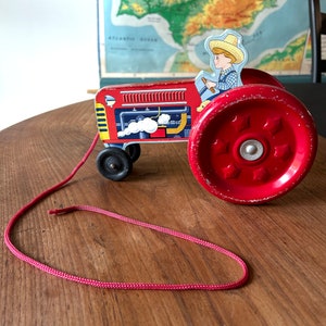 May include: Red toy tractor with a pull string, a cartoon farmer figure, and large wheels. The tractor has a detailed engine and a white smoke plume.