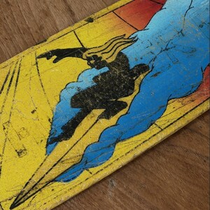 May include: A vintage skateboard deck featuring a black silhouette of a figure surfing a blue wave against a yellow and red background. The deck has a distressed, worn appearance.