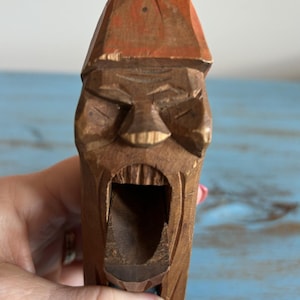 May include: A wooden nutcracker carved in the shape of a man's face with a wide open mouth. The nutcracker is painted in shades of brown and orange.