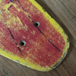 May include: A vintage skateboard deck with a distressed, worn appearance. The deck features a red and yellow color scheme with black accents and two circular holes. The surface shows signs of wear and tear, suggesting it has been used.