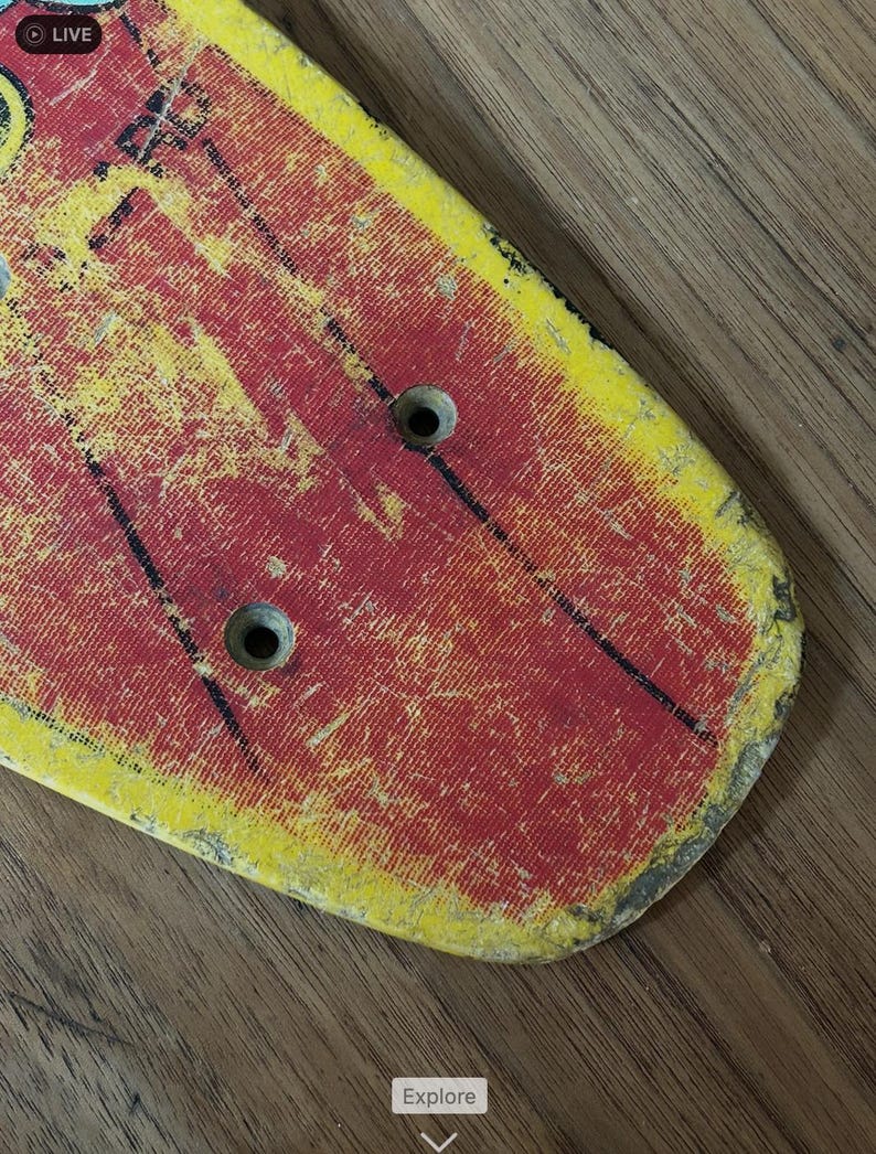 May include: A vintage skateboard deck with a distressed, worn appearance. The deck features a red and yellow color scheme with black accents and two circular holes. The surface shows signs of wear and tear, suggesting it has been used.