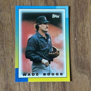 May include: Topps baseball card featuring Wade Boggs in a navy blue jacket and baseball cap. The card has a blue border and the player's name is printed in bold white letters. The background shows a stadium.