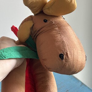 May include: A brown plush moose with a green and red ribbon around its neck. The moose has black eyes and a brown nose.