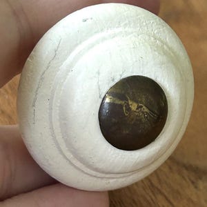 Peut inclure: Un bouton de meuble rond, blanc cass&eacute;, avec un centre brun fonc&eacute;. Le bouton pr&eacute;sente un design stri&eacute; et un centre de couleur laiton. Le bouton est tenu dans la main d'une personne.