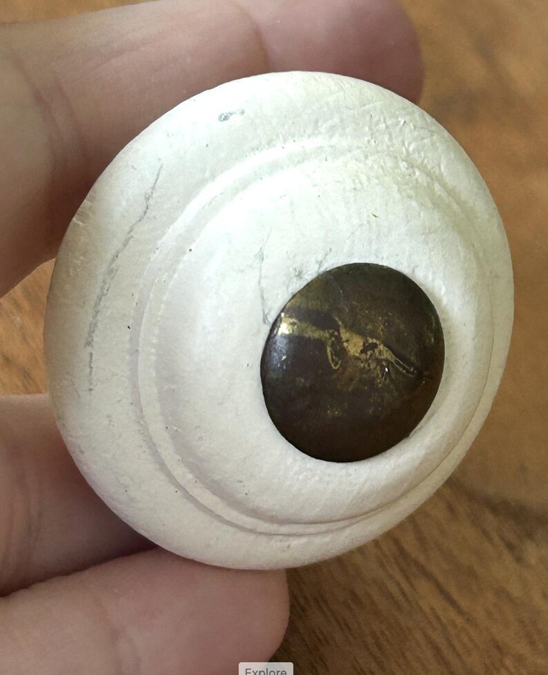 Peut inclure: Un bouton de meuble rond, blanc cass&eacute;, avec un centre brun fonc&eacute;. Le bouton pr&eacute;sente un design stri&eacute; et un centre de couleur laiton. Le bouton est tenu dans la main d'une personne.