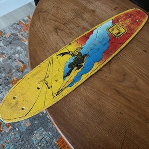 May include: Vintage yellow skateboard with a graphic of a surfer riding a wave. The board has a red and blue design with the words "HANG TEN" in a yellow square. The skateboard is on a wooden table.