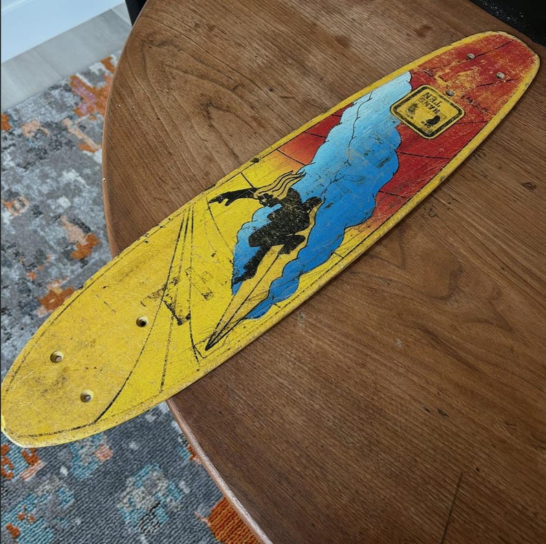 May include: Vintage yellow skateboard with a graphic of a surfer riding a wave. The board has a red and blue design with the words "HANG TEN" in a yellow square. The skateboard is on a wooden table.