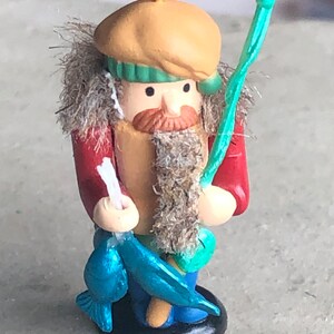 Puede incluir: Una pequeña figura de madera pintada a mano de un pescador que lleva un sombrero marrón con una banda verde, una chaqueta roja y una camisa azul. Lleva una caña de pescar y un pez azul.