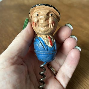 Pode incluir: Um saca-rolhas vintage com uma cabeça de madeira pintada de um homem vestindo uma jaqueta azul e um chapéu preto. O saca-rolhas é feito de metal e tem um design em espiral.