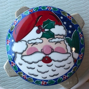 Puede incluir: Una caja de cerámica para guardar pequeños objetos navideños con un borde azul y verde. La tapa tiene un dibujo de Papá Noel con un gorro rojo, barba blanca y nariz rosa.