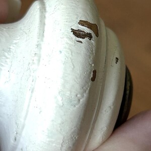 Peut inclure: Gros plan sur un bouton d&eacute;coratif blanc avec une finition vieillie. Le bouton a une forme arrondie avec des rainures verticales et des zones visibles de bois apparent. Le bouton est tenu par une main.