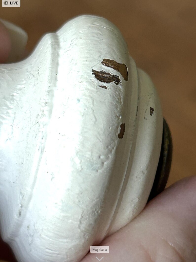 Peut inclure: Gros plan sur un bouton d&eacute;coratif blanc avec une finition vieillie. Le bouton a une forme arrondie avec des rainures verticales et des zones visibles de bois apparent. Le bouton est tenu par une main.